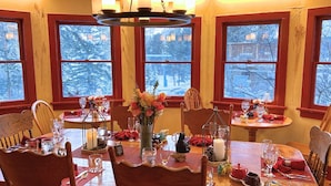A dining area with a wooden table set for four, surrounded by chairs. The room has large windows with a view of snow-covered trees. - Chiming Bells Suite at RiverSong Inn in Estes Park - Riverside in Rocky Mountain National Park (Estes Park)