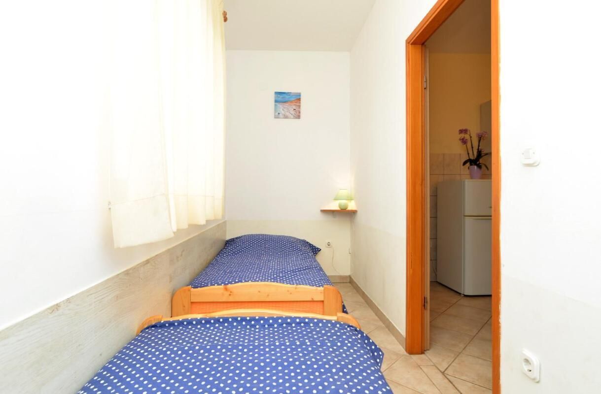Una habitación pequeña y estrecha con dos camas individuales, un pequeño estante con una planta y vistas a la cocina a través de una puerta abierta.