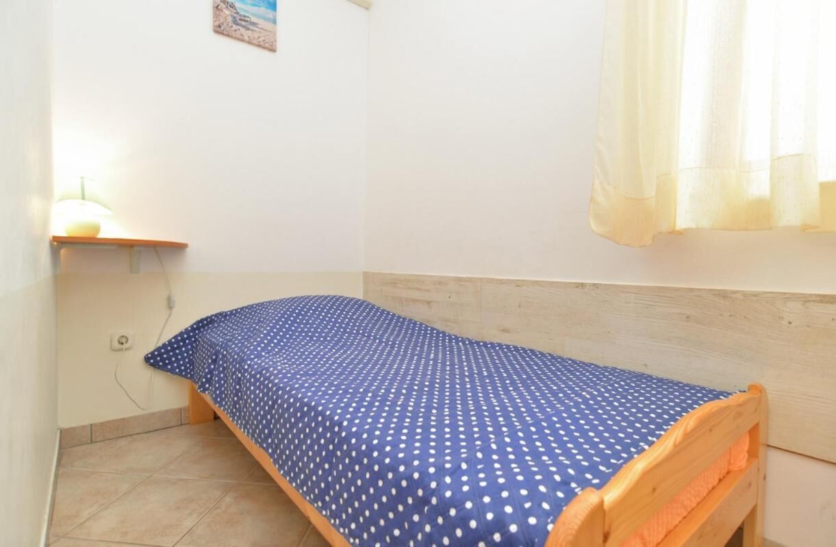 A small, single-bed room with a wooden headboard, a blue polka-dotted bedspread, a small shelf with a lamp, and a window with sheer curtains.