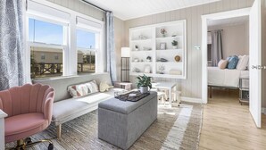 Una sala de estar acogedora con un sillón rosa, un otoman gris y un sofá blanco. Hay un aparador blanco con adornos y una lámpara.