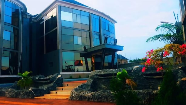 A modern building with a curved glass facade, a covered entrance, and a landscaped area with rocks and tropical plants. - One World Luxury Hotel (Enugu)