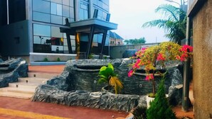 A modern building with a glass facade, a landscaped area with rocks and plants, and a small structure with a roof. - One World Luxury Hotel (Enugu)