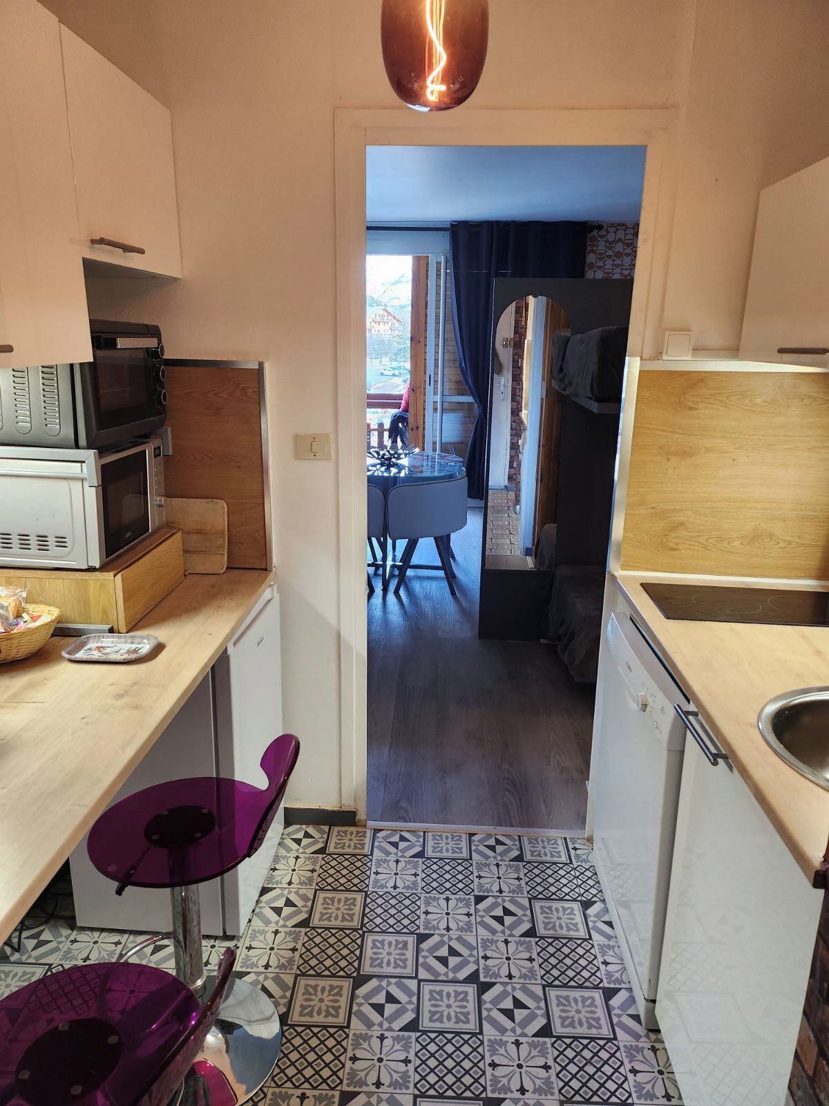 A compact kitchen with a microwave, toaster, and a small sink. A purple chair is visible, and the floor has patterned tiles.