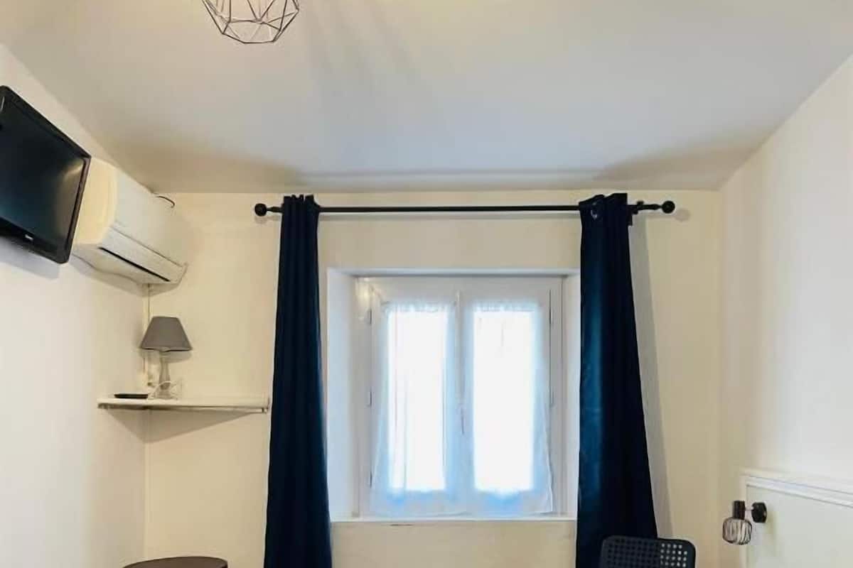 Quarto com cama, televisão na parede, janela com cortinas, uma mesinha e uma cadeira.