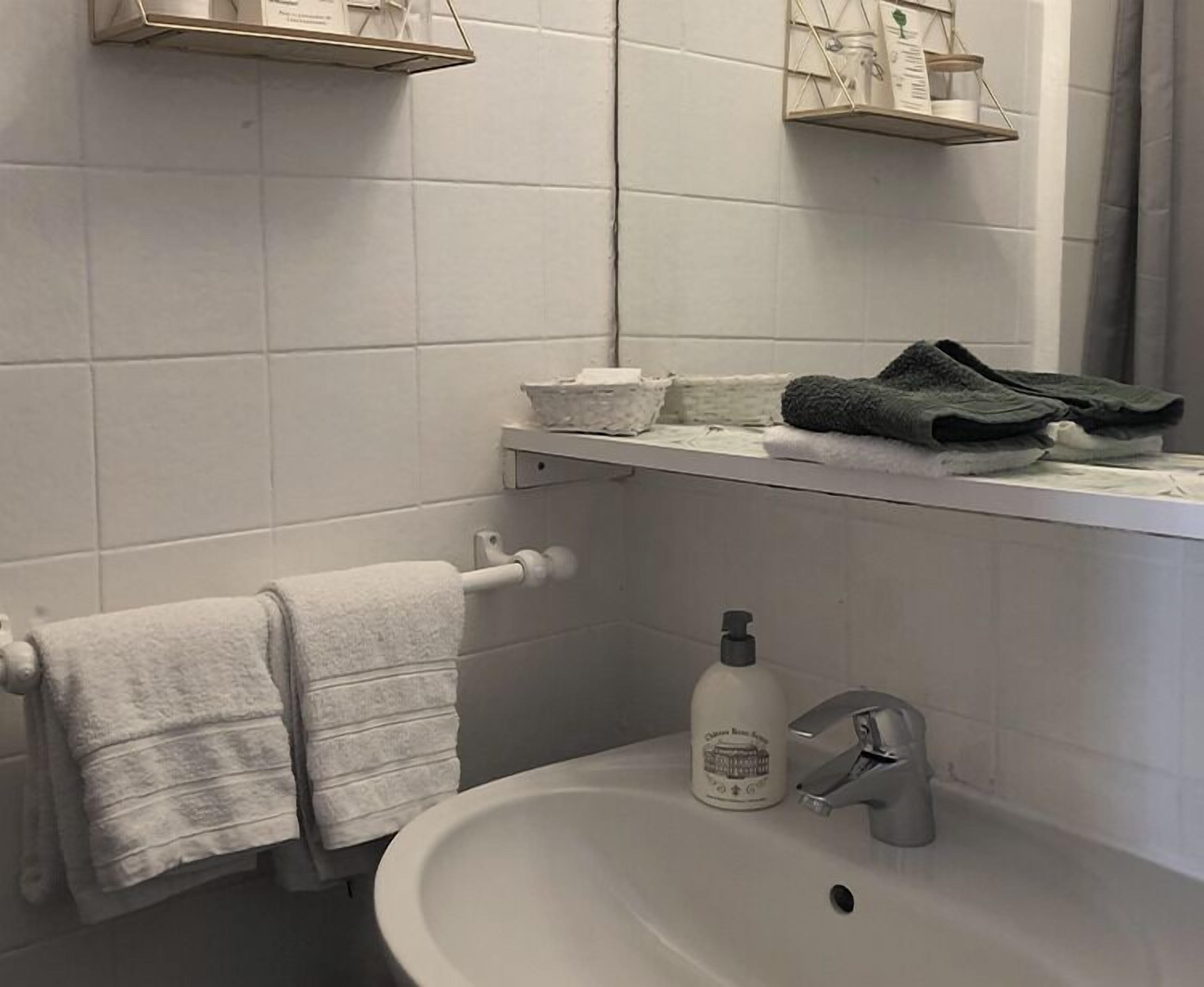 Un baño con un lavamanos blanco, un perchero con una toalla doblada, una botella de jabón para manos y un par de zapatillas en un estante.