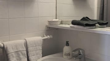 Un baño con un lavamanos blanco, un perchero con una toalla doblada, una botella de jabón para manos y un par de zapatillas en un estante.