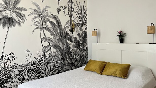 Une chambre à coucher ornée d’une grande fresque murale aux motifs tropicaux, avec un lit blanc et un oreiller de couleur moutarde, ainsi qu’une tête de lit blanche.