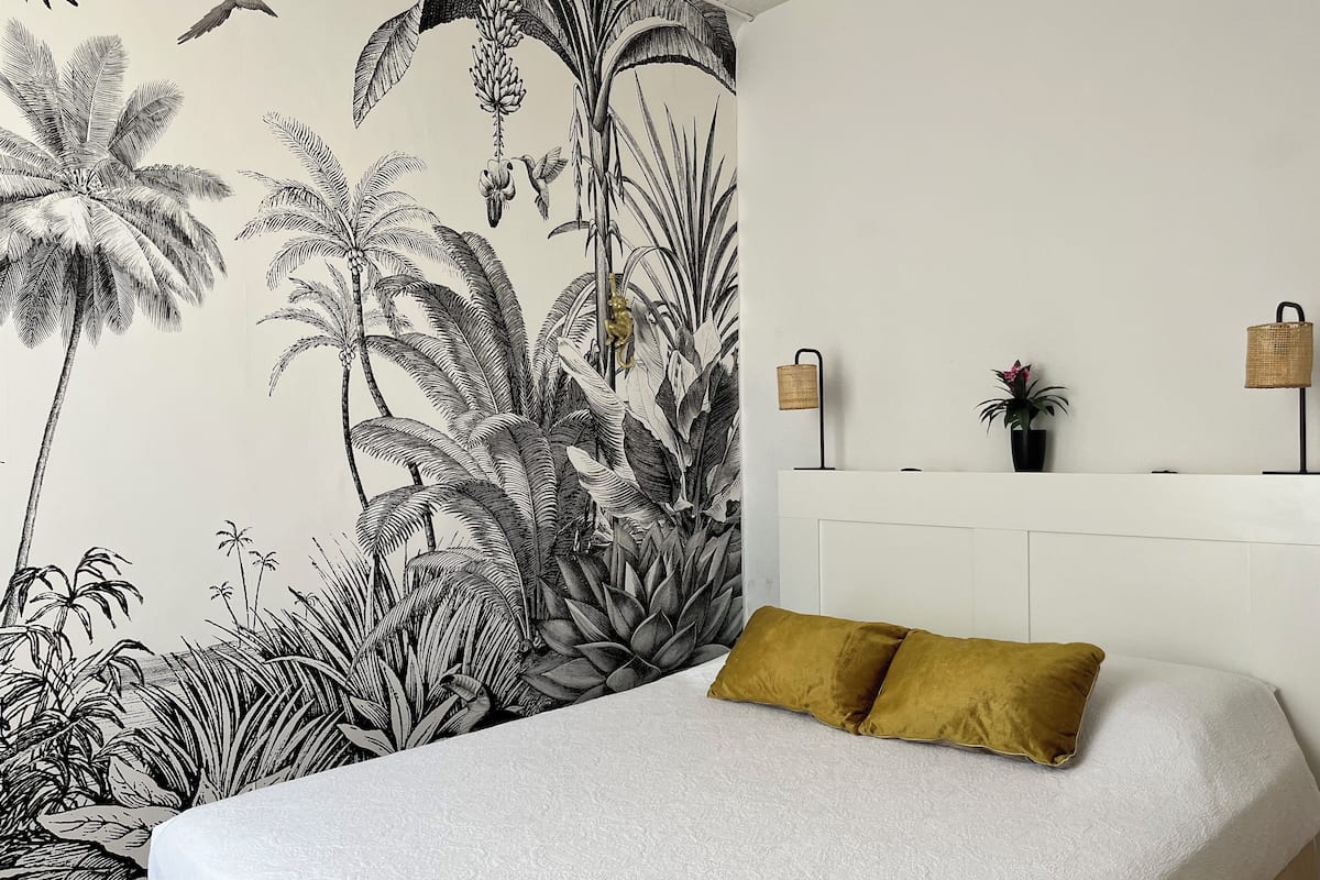 Quarto com um grande mural tropical, cama branca com travesseiro amarelo mostarda e cabeceira branca.
