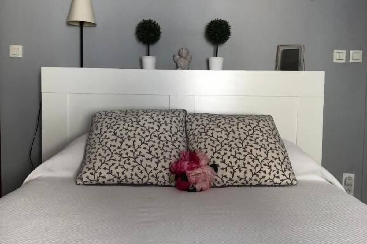 Cama bem arrumada com almofadas estampadas e um arranjo floral decorativo, cabeceira branca e luminárias fixadas na parede.