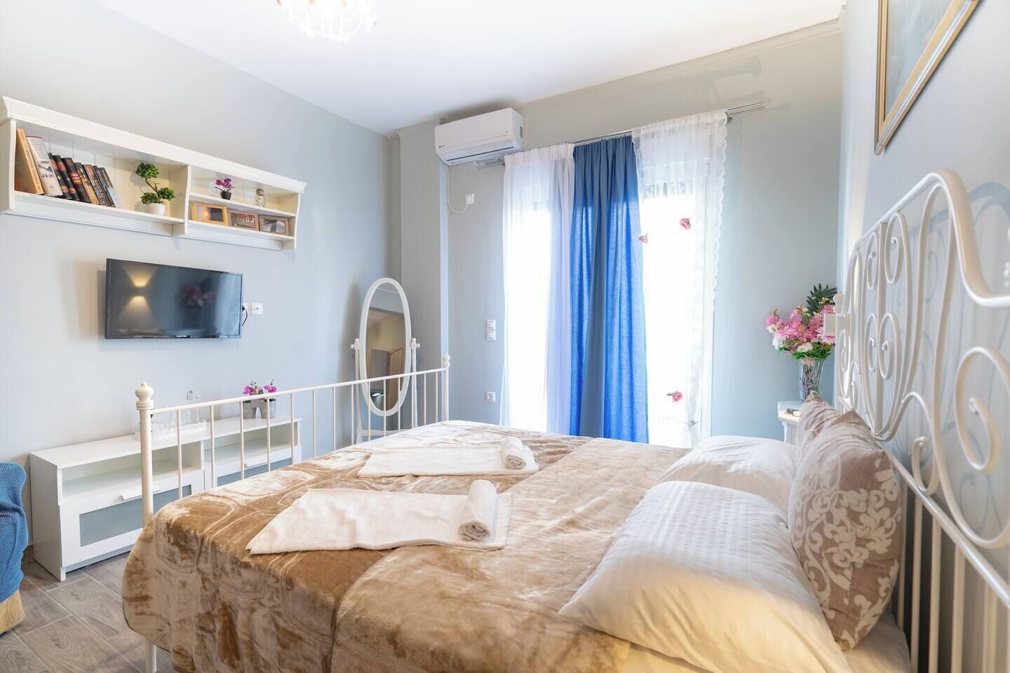A bedroom with a bed, a TV, a mirror, a shelf with books, and a window with curtains.