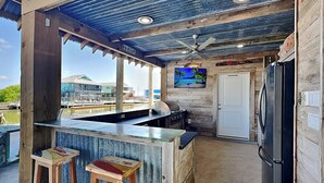 A rustic outdoor kitchen with a bar area, wooden stools, a refrigerator, and a television displaying a beach scene.