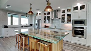 A modern kitchen with a central island, wooden stools, white cabinetry, and stainless steel appliances.