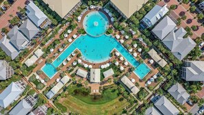 An aerial view of a resort with multiple swimming pools, arranged in a central circular pattern, surrounded by lounge areas and palm trees.