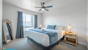 A bedroom with a bed, a ceiling fan, a bedside table with a lamp, a window with curtains, and a mirror. - Newly Built Retreat | Comfort & Ski Access (American Fork)