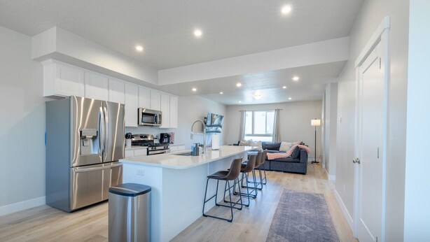 A modern kitchen with stainless steel appliances, a central island with bar stools, and a cozy seating area with a sofa and lamp. - Newly Built Retreat | Comfort & Ski Access (American Fork)