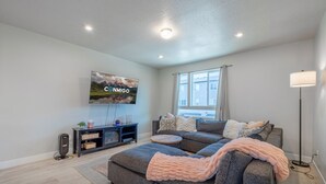 A modern living room with a sectional sofa, a flat-screen TV mounted on the wall, a floor lamp, and a window with curtains. - Newly Built Retreat | Comfort & Ski Access (American Fork)