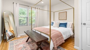 Une chambre à coucher avec un lit à baldaquin, un banc, un grand miroir et une vue sur la verdure par la fenêtre.