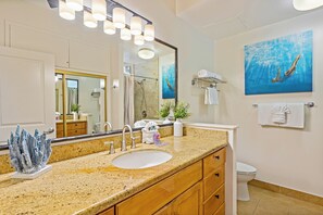 A bathroom with a large mirror, a sink, and a toilet. There is a painting of a diver on the wall and a plant on the counter. - Gorgeous Napili Shores G258 Newly remodeled Studio Condo (lahaina)