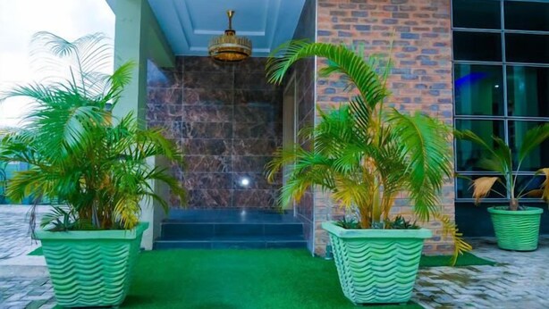 A building entrance with a green artificial grass pathway, two large potted plants, a brick wall, and a glass door. - E- Exclusive Hotel  (Ibadan)