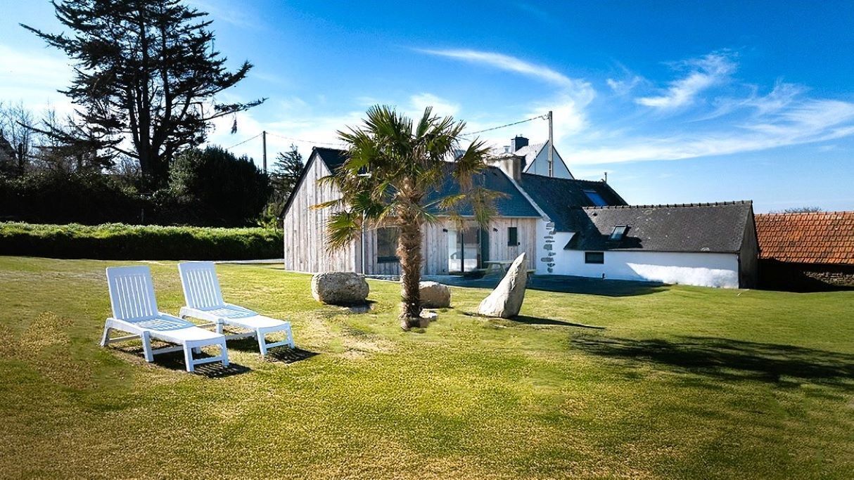 Een wit gebouw met een schuin dak, een palmboom en twee witte lounge stoelen op een gazon.