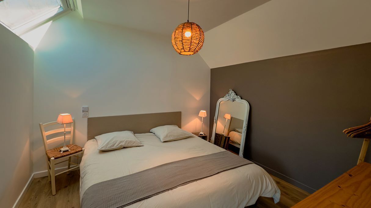 A bedroom with a bed, a chair, a bedside table, a mirror, and a hanging light fixture.