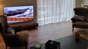 Ein Wohnzimmer mit Fernseher, zwei Sesseln, einem Couchtisch und einem großen Fenster mit durchscheinenden Vorhängen.