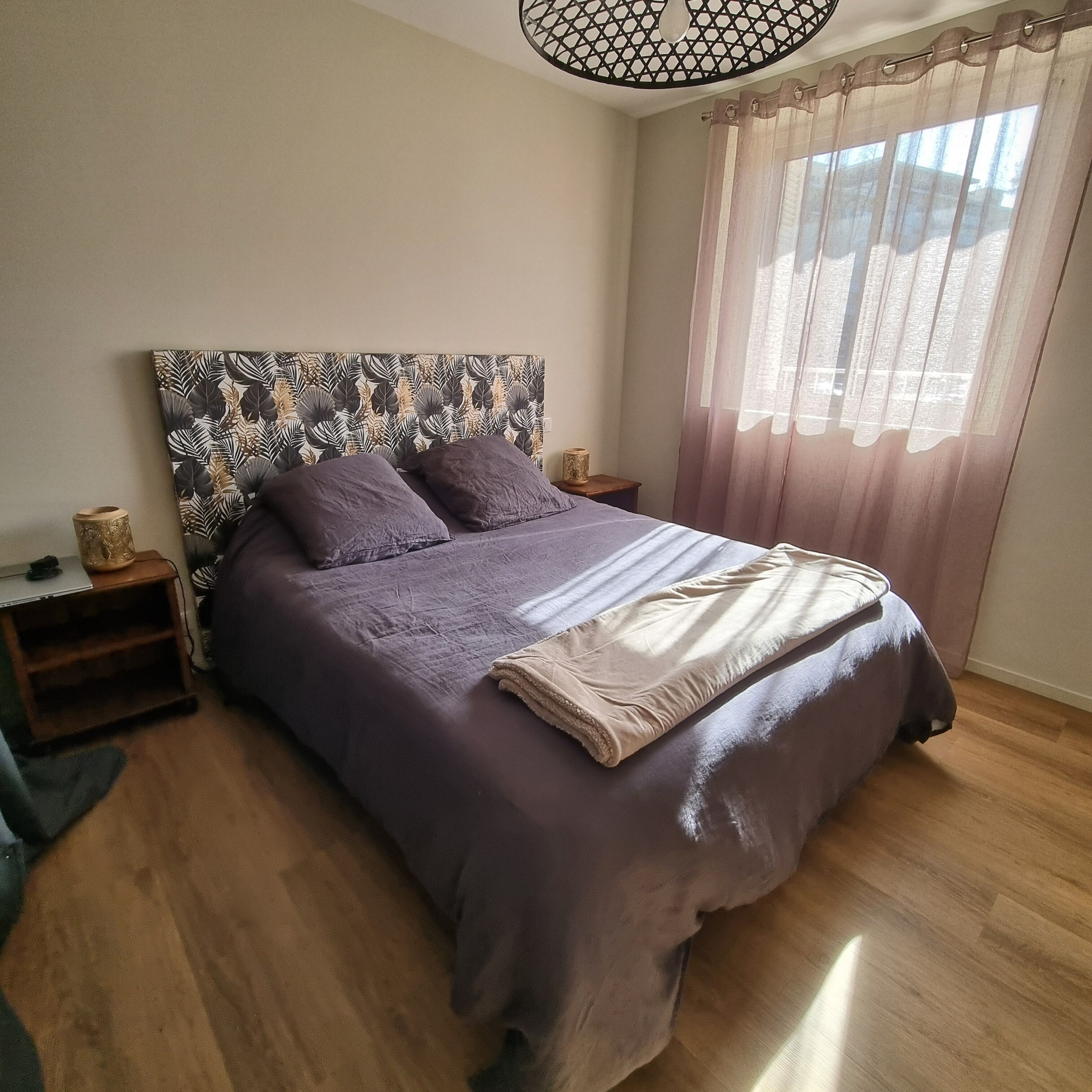 A bedroom with a bed, a headboard with a leaf pattern, a bedside table, a lamp, and a window with curtains.