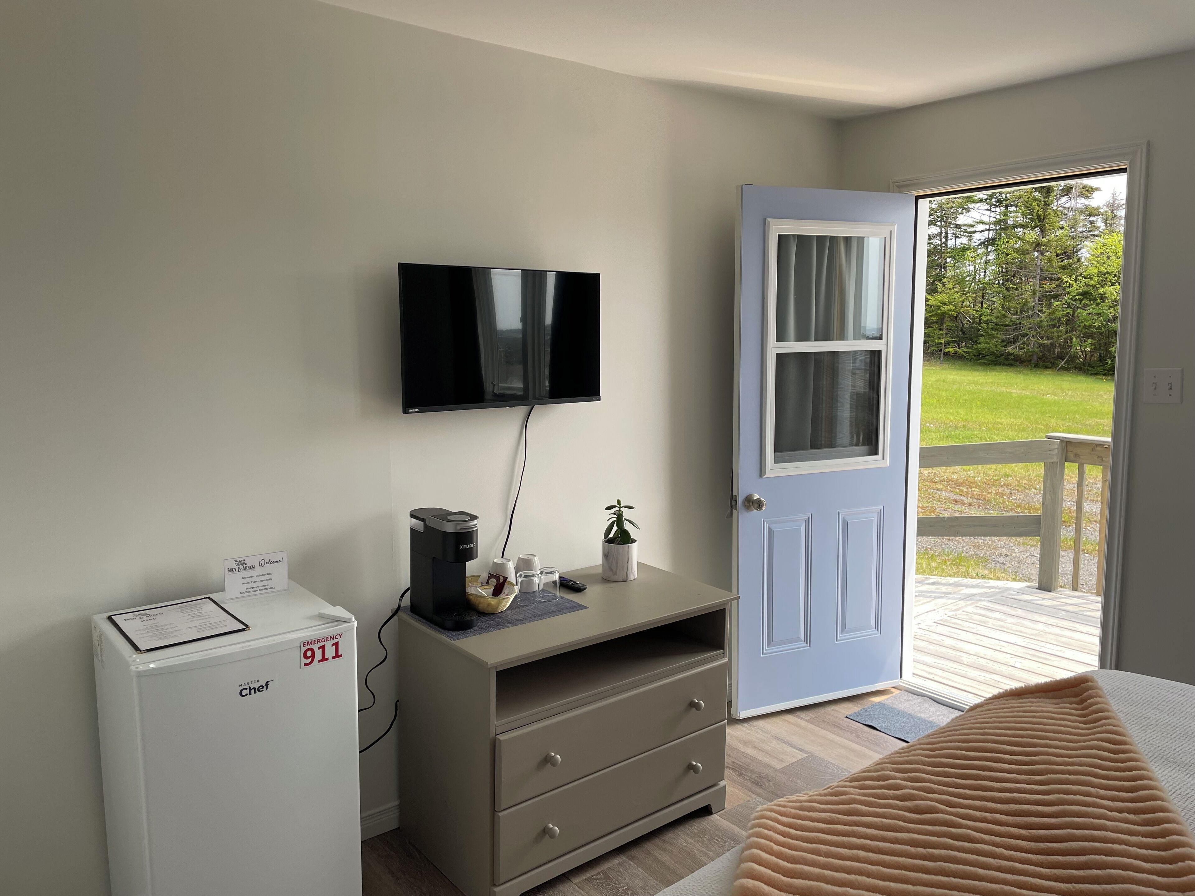 Hotelkamer met een flatscreen televisie aan de muur, een minibar, een klein tafeltje met een plant, een deur naar een balkon en een bed met een beige deken.