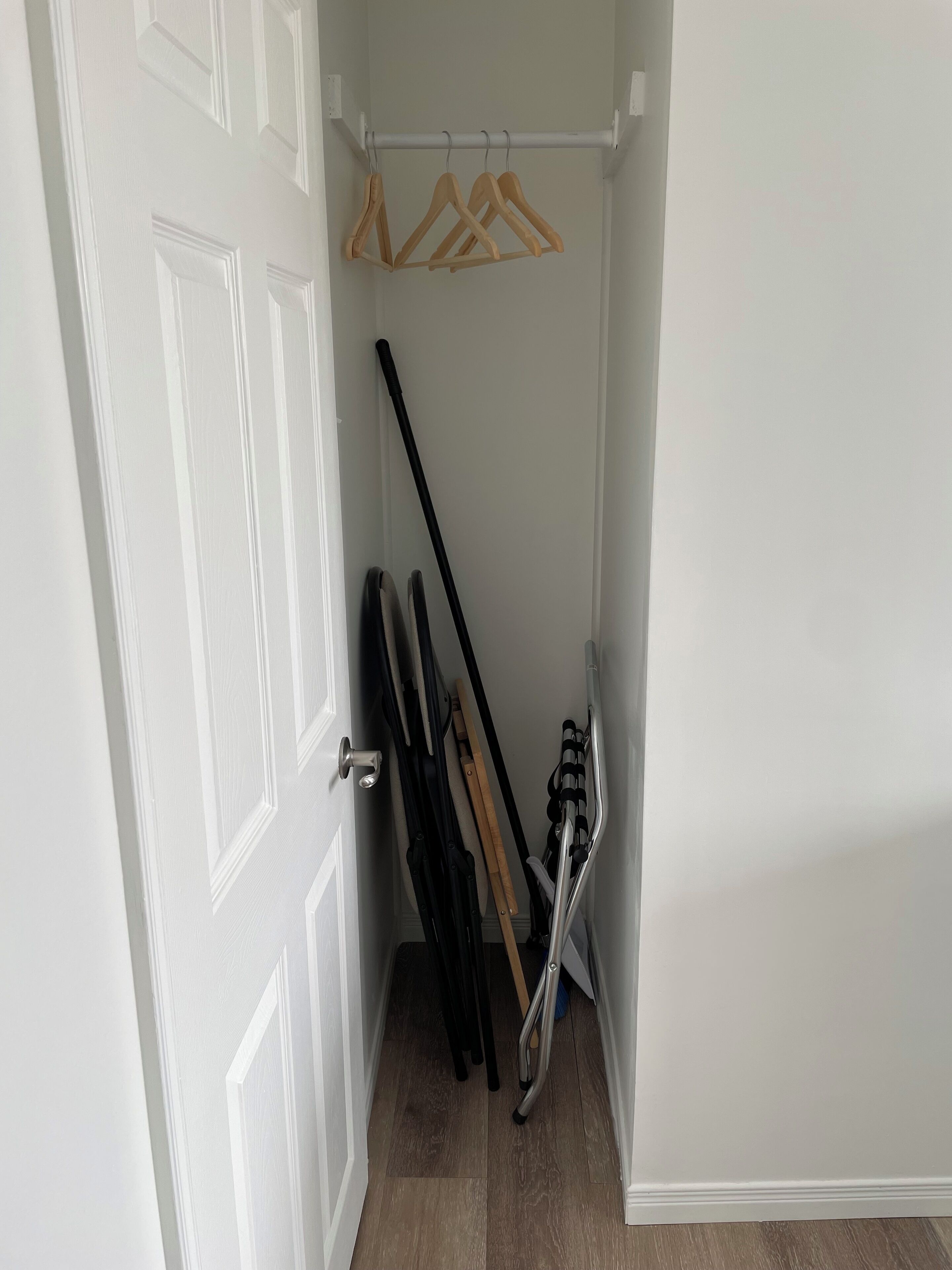 Un pasillo estrecho con una puerta blanca, un perchero con perchas de madera y una silla plegable de metal.