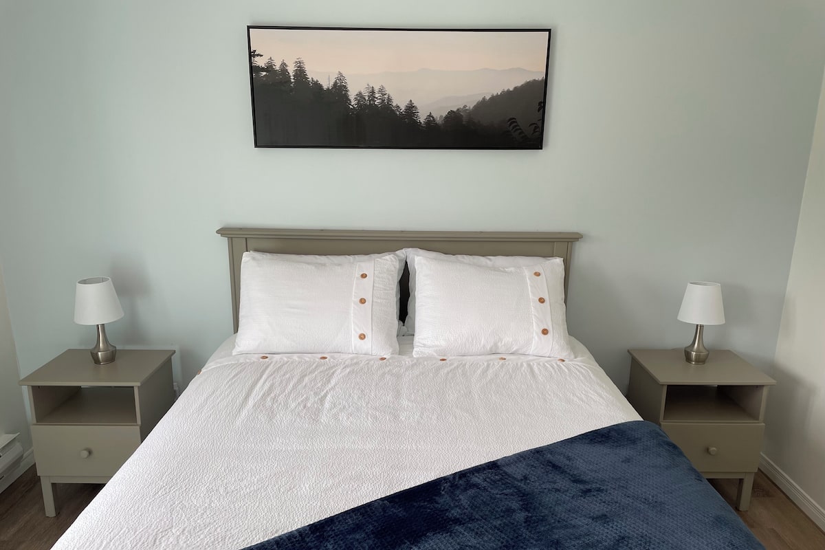Una cama bien hecha con ropa blanca y dos almohadas, flanqueada por dos mesitas de noche con lámparas, en una habitación con un cuadro de un paisaje de montaña en la pared.