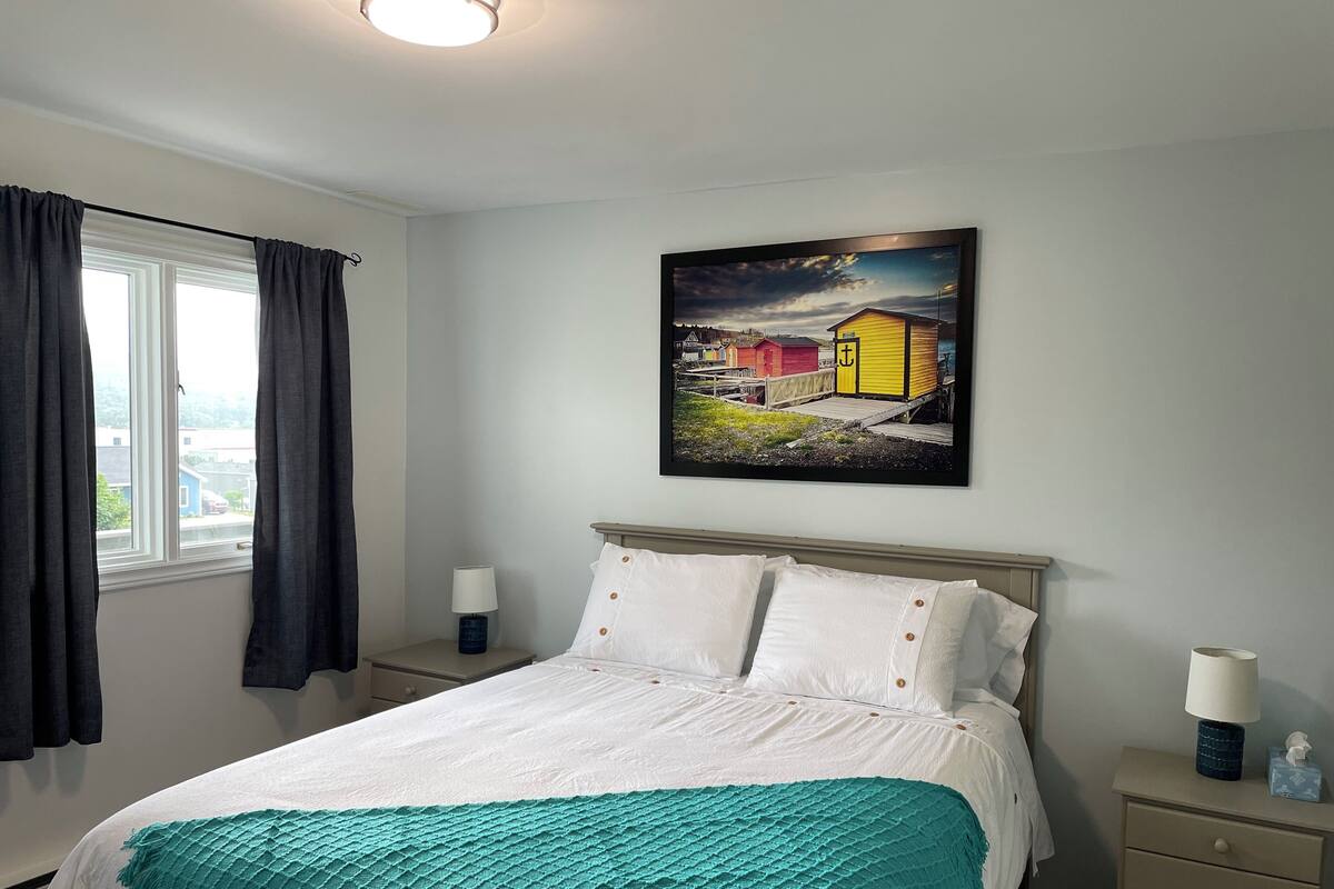 Un dormitorio con cama, mesita de noche, lámpara, un cuadro y ventana con cortinas.