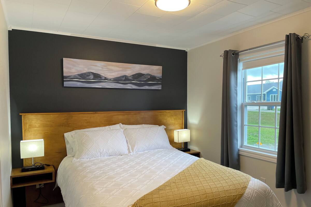 Una cama bien hecha con cabecera, dos lámparas de noche, una mesita de noche con lámpara, un cuadro en la pared y una ventana con cortinas.