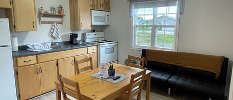 Cocina compacta con armarios de madera, un microondas y un lavamanos. Hay una mesa de comedor con sillas y un sofá negro.