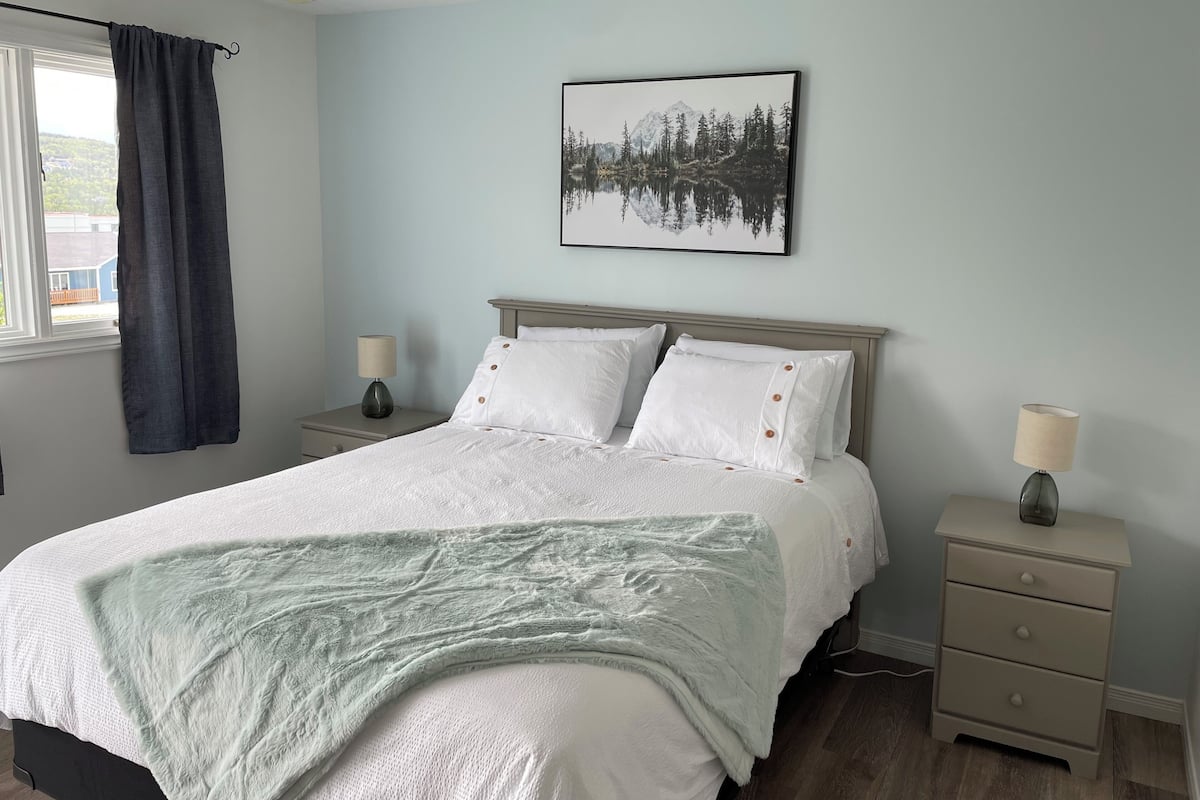 Un dormitorio con cama, dos mesitas de noche, una ventana con cortinas, un cuadro en la pared y una lámpara de noche.