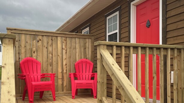 A wooden deck with two red chairs and a wooden staircase leading to a red door. - Buoy & Arrow Restaurant and Suites (Rocky Harbour)