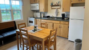A compact kitchen with wooden cabinets, a microwave, a stove, and a refrigerator. There is a dining table with chairs and a bench. A window provides a view of the outdoors. - Buoy & Arrow Restaurant and Suites (Rocky Harbour)