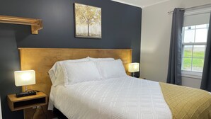 A neatly made bed with white bedding, two bedside lamps, a wooden headboard, a wall-mounted painting, and a window with curtains. - Buoy & Arrow Restaurant and Suites (Rocky Harbour)