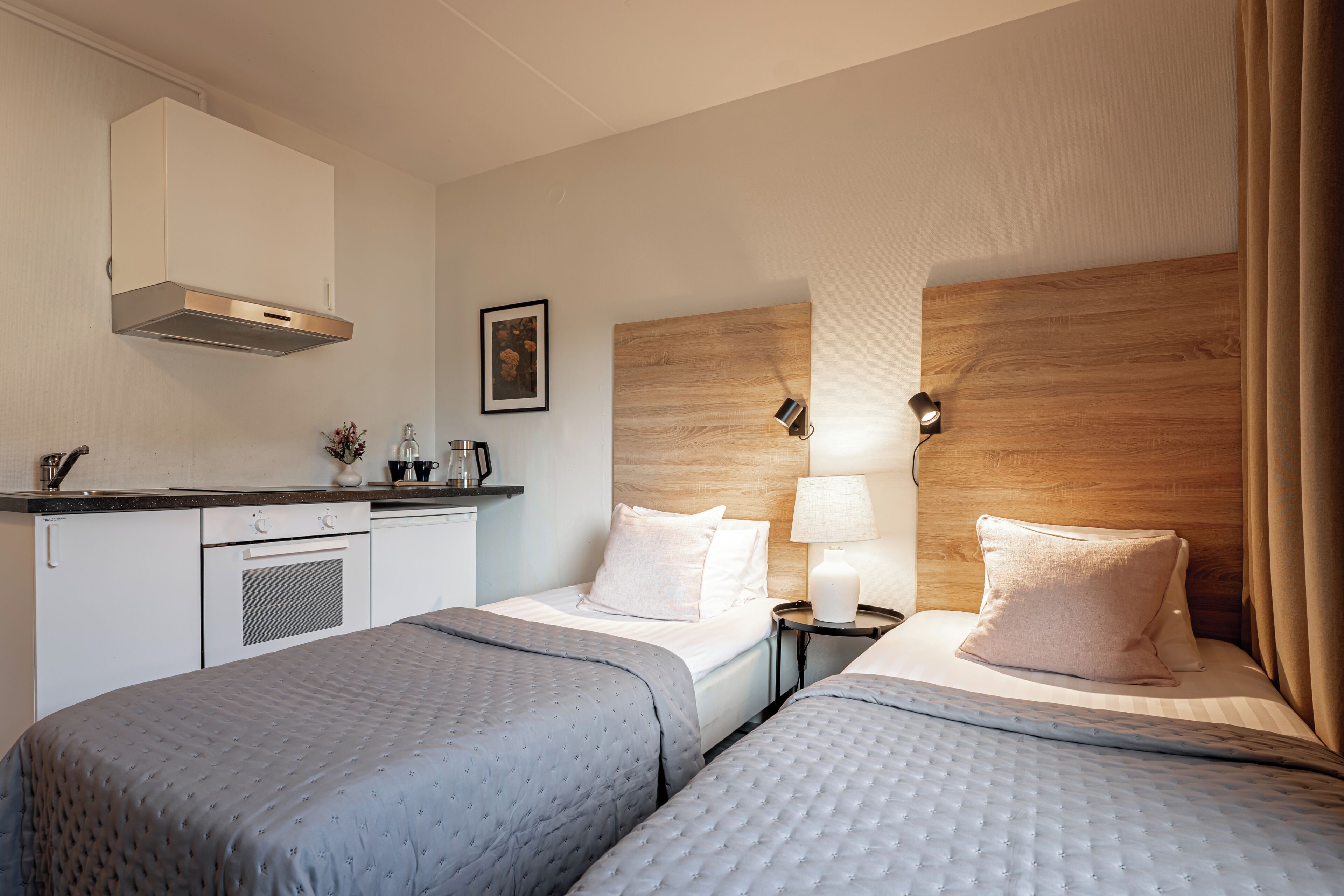 A hotel room with two beds, a kitchenette, a wooden headboard, and a nightstand with a lamp.
