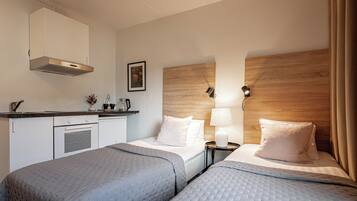 A hotel room with two beds, a kitchenette, a wooden headboard, and a nightstand with a lamp.