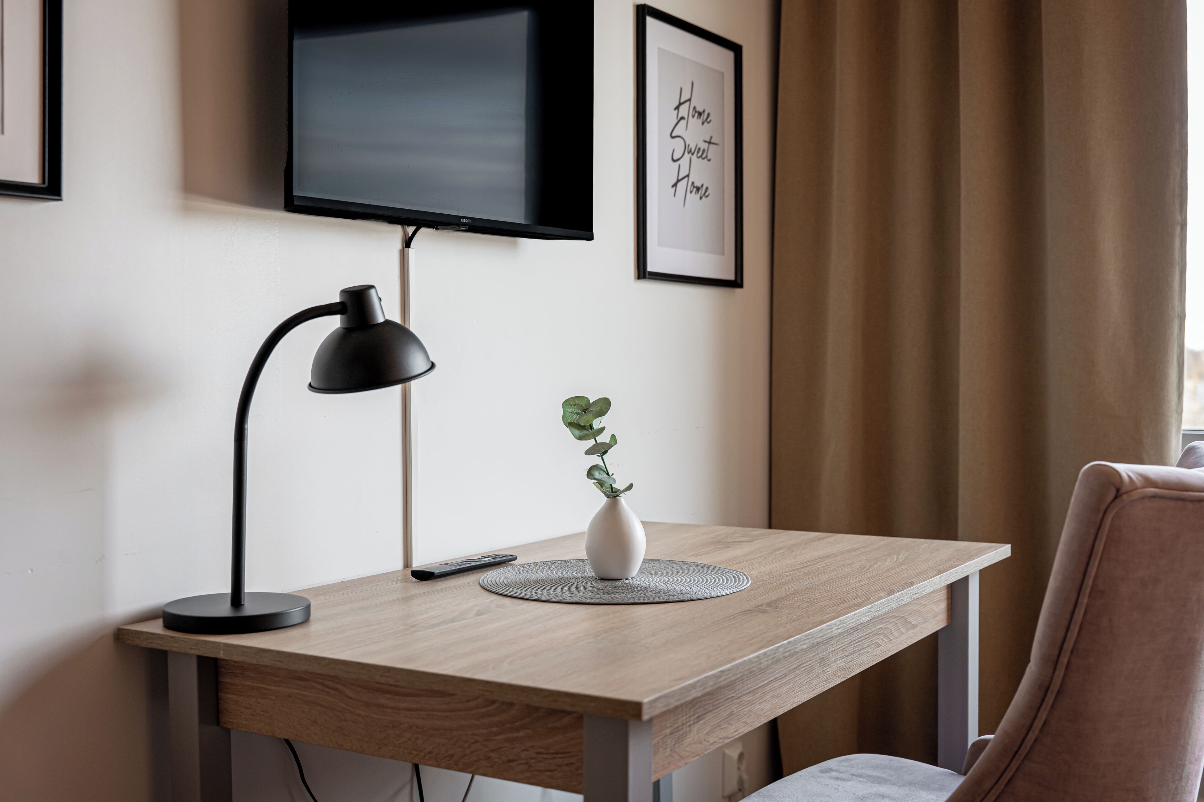 Ett skrivbord i trä med en svart skrivbordslampa, en fjärrkontroll och en vit vas med en växt. En tv är monterad på väggen ovanför skrivbordet.