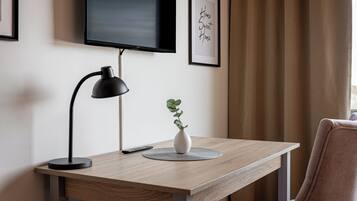 Ett skrivbord i trä med en svart skrivbordslampa, en fjärrkontroll och en vit vas med en växt. En tv är monterad på väggen ovanför skrivbordet.