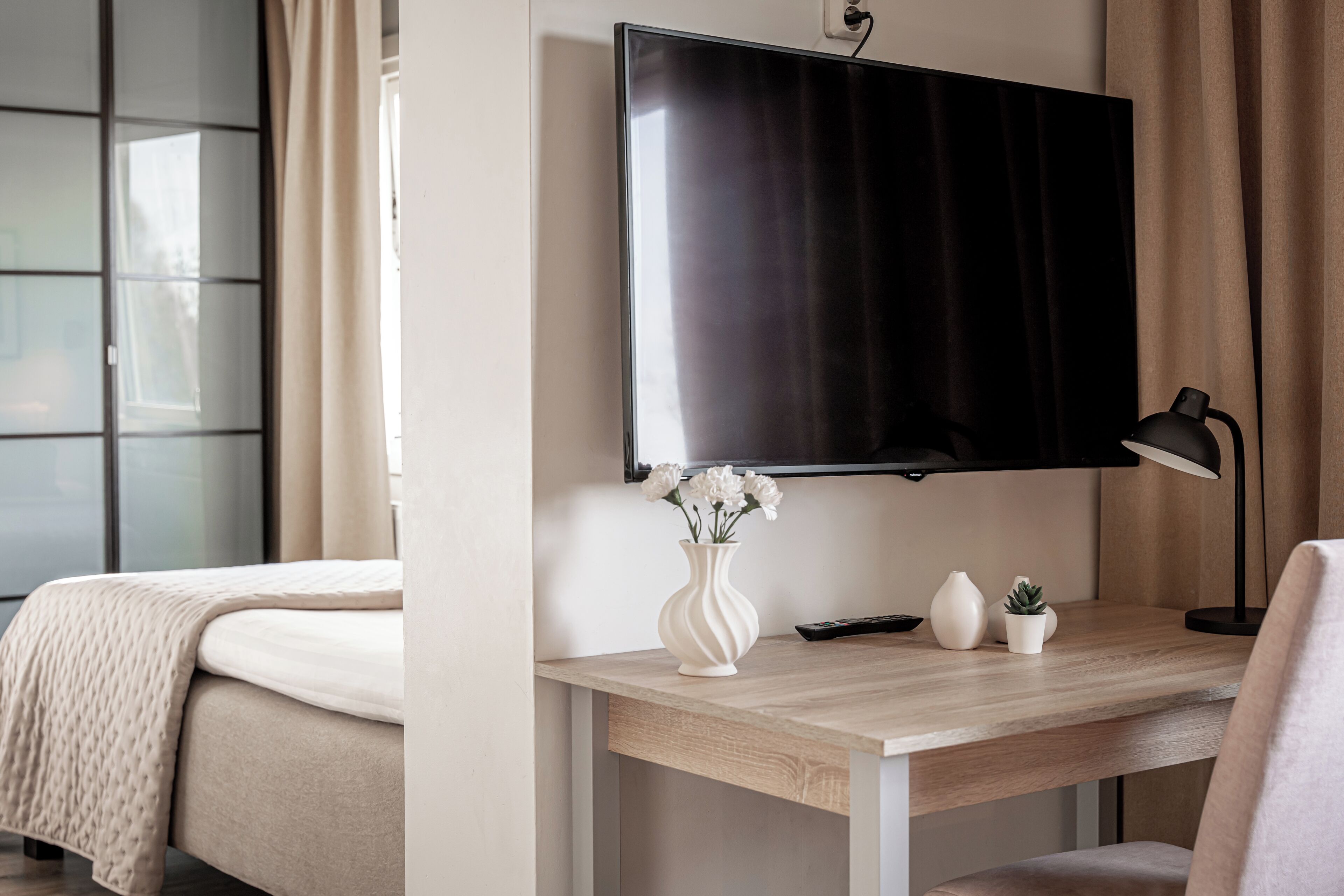 A hotel room with a bed, a desk with a TV, a lamp, and a vase with flowers.
