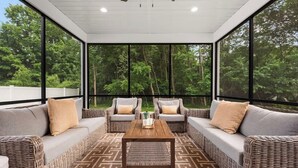 A covered outdoor seating area with wicker furniture, a central coffee table, and large windows offering a view of a forest.
