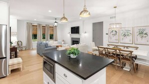 A modern kitchen with a central island, dining area with wooden chairs, and a living room with a sofa and fireplace.
