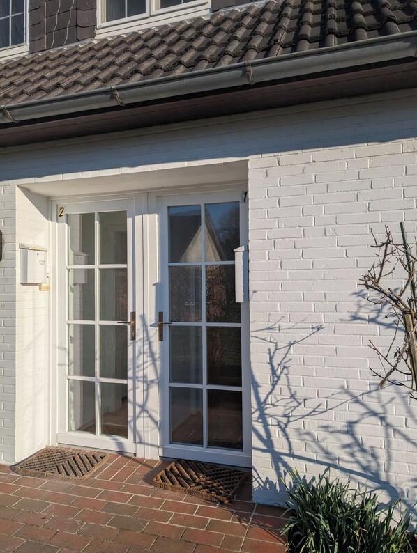 A white brick building with a tiled roof, a white door with glass panels, and a small doormat. - ***SANDLOFT*** (Sankt Peter-Ording)
