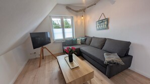 A cozy living room with a grey sofa, a wooden coffee table with a vase of flowers, a flat-screen TV on a wooden stand, and a window with blinds. - ***SANDLOFT*** (Sankt Peter-Ording)