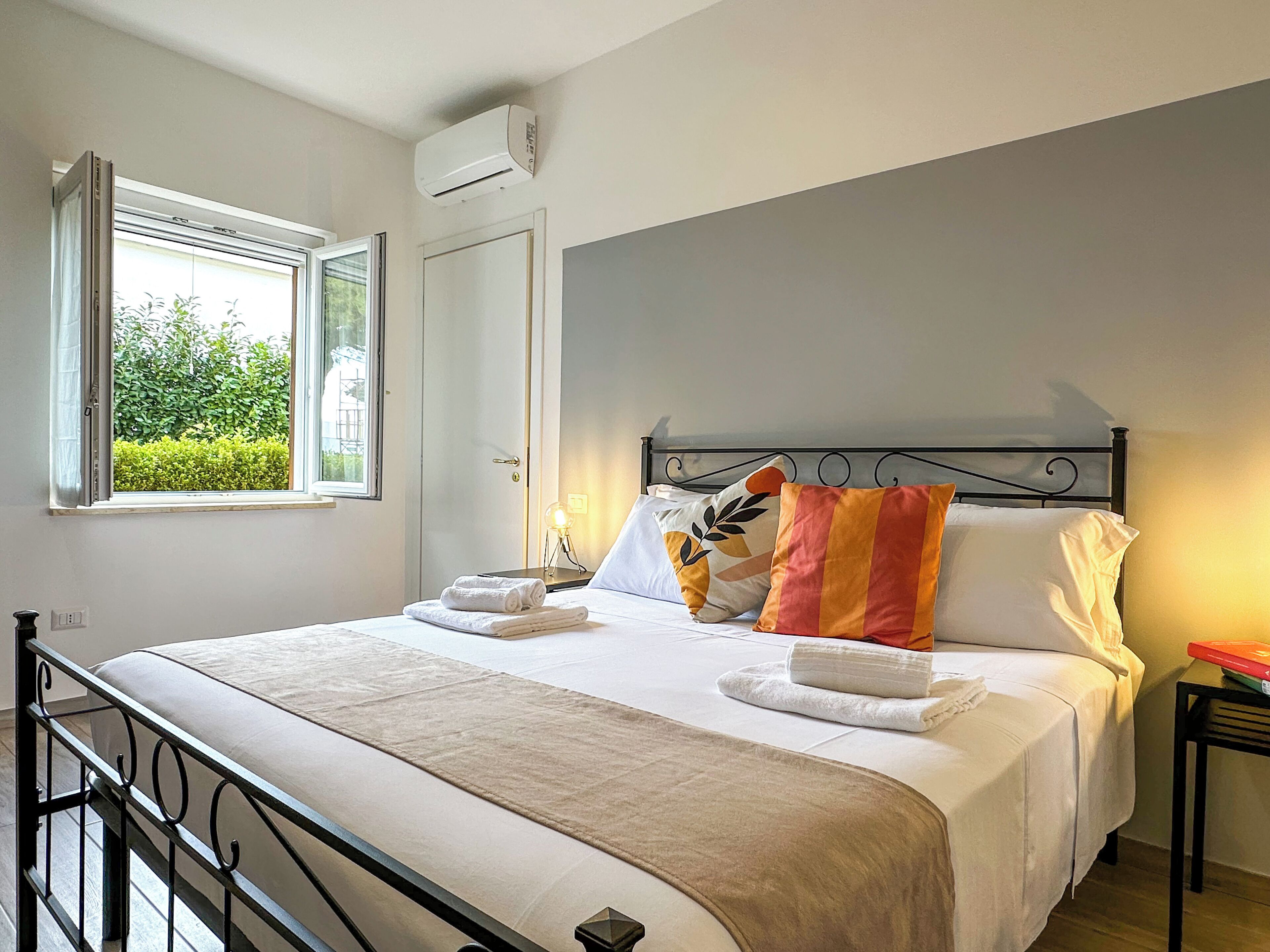 Un letto rifatto con lenzuola bianche e numerosi cuscini, un comodino con una lampada, e una finestra con vista sul verde.