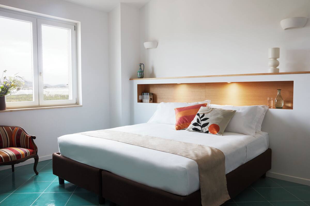 A neatly made bed with a wooden headboard, a window with a view, and a chair with a striped cushion.