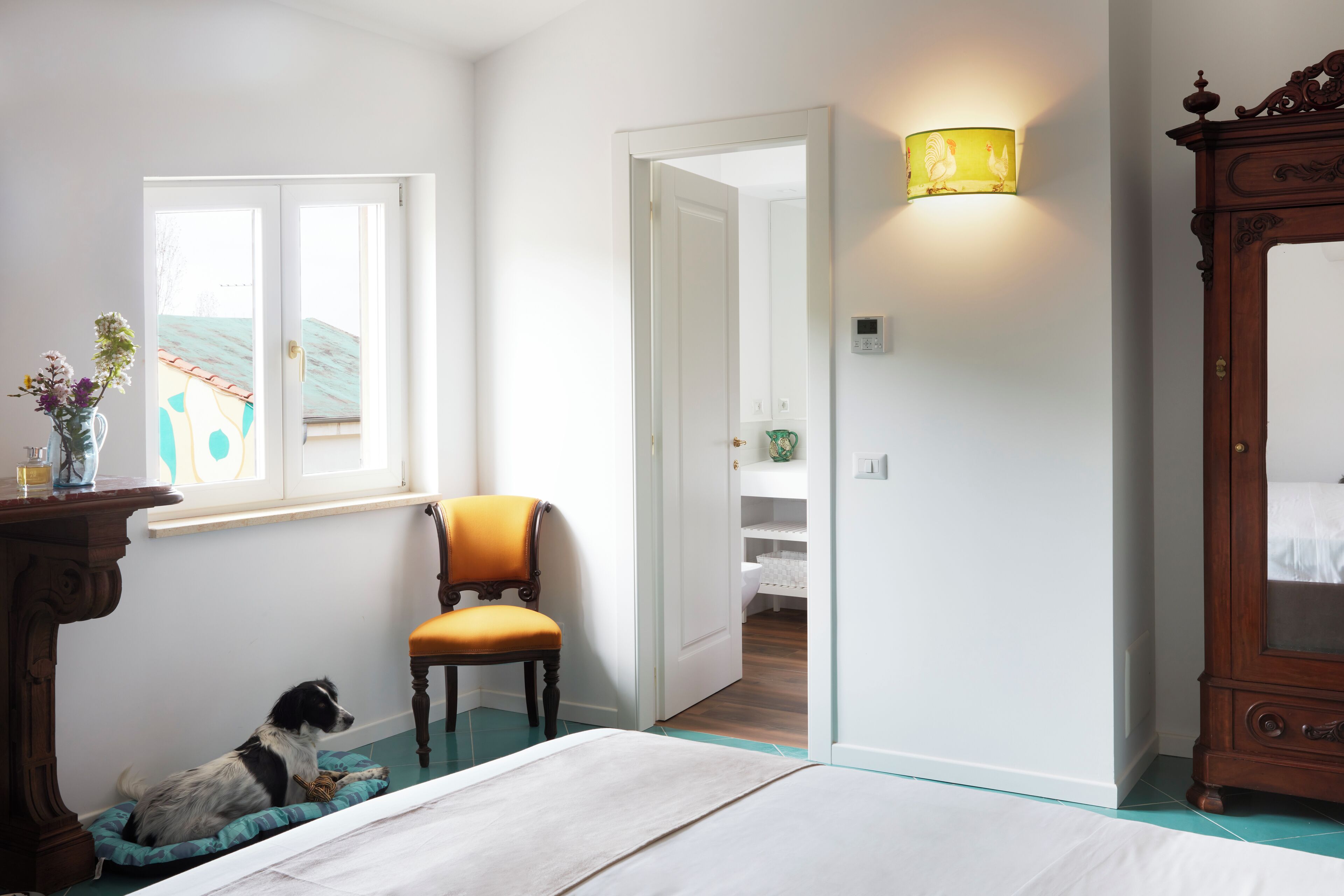 A bedroom with a bed, a chair, a dog, a dresser, and a lamp.