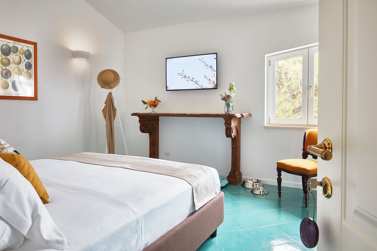 A bedroom with a bed, a wooden table with a vase, a TV, a window, and a door.